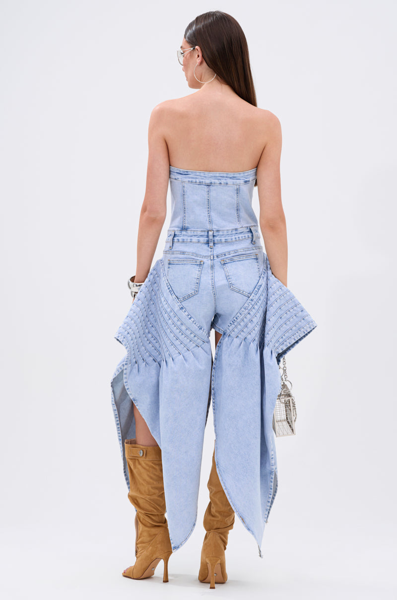 A woman stands facing away, wearing the NO ONE LIKE ME DENIM ROMPER, paired with tan knee-high boots and a small handbag. Her straight brown hair is down.