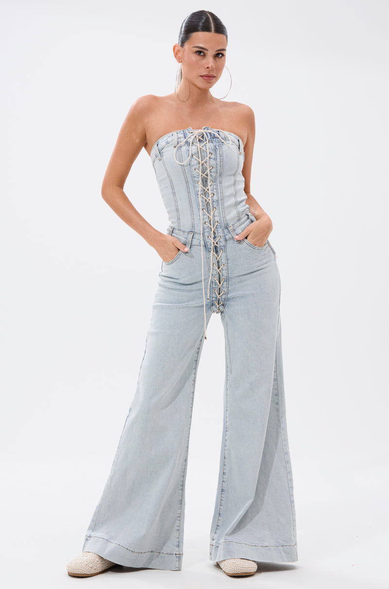 A woman poses confidently against a white background, hands in pockets, wearing the ALL IN THE DETAILS DENIM LACE UP JUMPSUIT—a light-wash denim piece featuring lace-up details and flared pant legs. Her hair is tied back.