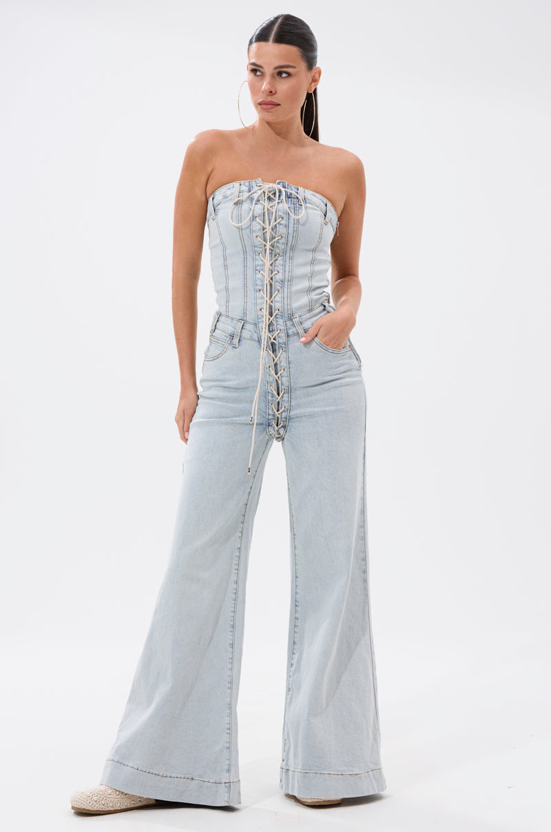 A woman models the ALL IN THE DETAILS DENIM LACE UP JUMPSUIT, featuring a light wash, lace-up corset bodice, wide-leg pants, and hoop earrings, standing with one hand in her pocket against a plain white background.