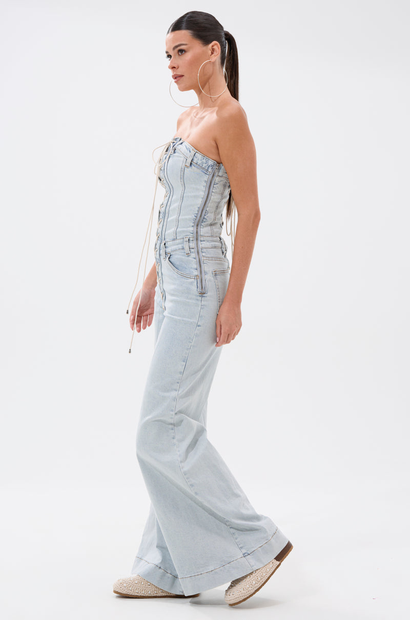 A woman in profile wears the ALL IN THE DETAILS DENIM LACE UP JUMPSUIT, a western-inspired, strapless piece with a lace-up front and flared legs. She styles her long hair in a sleek ponytail and completes the look with espadrille platform shoes.