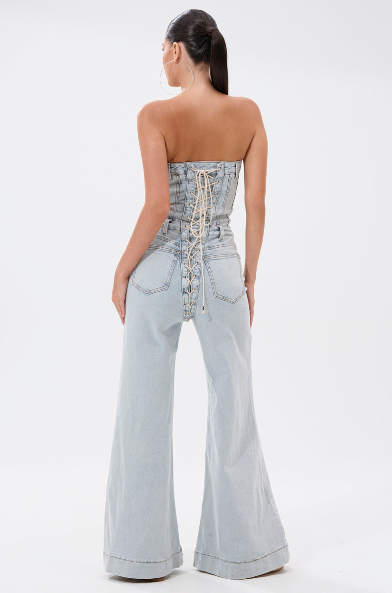 A woman with sleek dark hair in a ponytail faces away, wearing the ALL IN THE DETAILS DENIM LACE UP JUMPSUIT—a light-wash, high-waisted style with wide legs and lace-up back detailing.