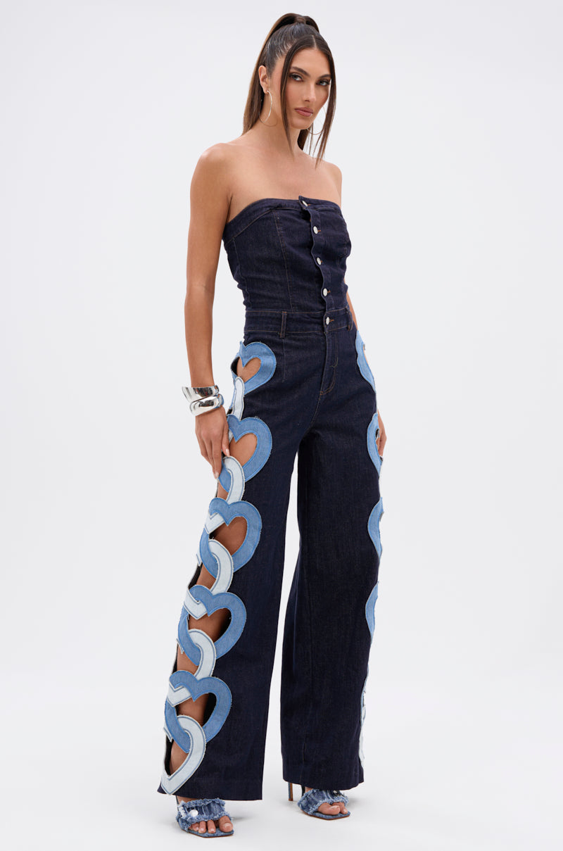 A woman models the HEART EYES FOR YOU DENIM JUMPSUIT, a strapless dark denim piece with front buttons and light blue heart cutouts on the sides. She completes the look with silver bracelets and high-heeled sandals.