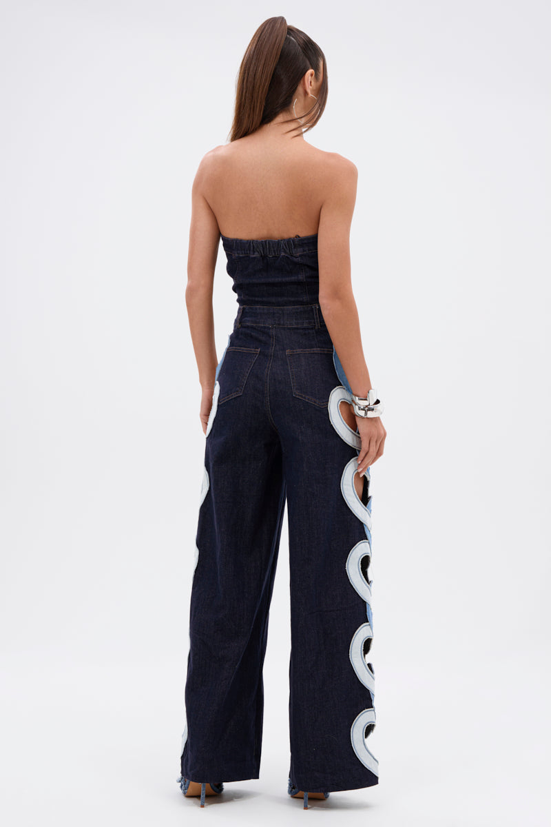 A woman with a high ponytail stands facing away, modeling the HEART EYES FOR YOU DENIM JUMPSUIT featuring dark denim, wide-leg jeans, and white wave-like patterns down the sides. She accessorizes with bracelets on one wrist.
