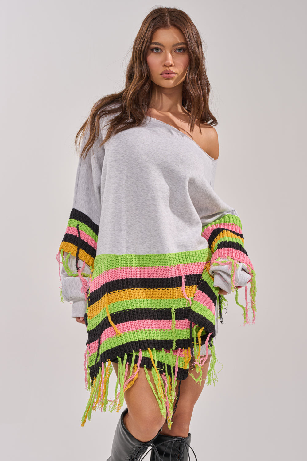 A woman models the MOST LOVED SWEATSHIRT MINI DRESS—an oversized crochet sweater dress with multicolored stripes and fringe—paired with black knee-high boots, standing against a plain background. 0