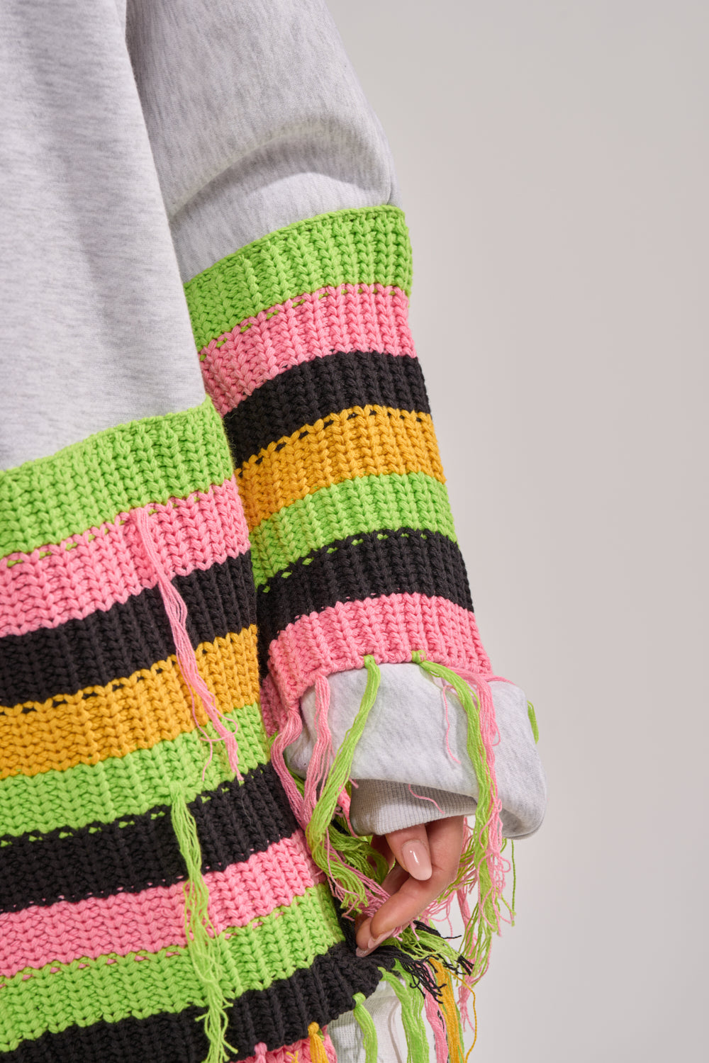 Close-up of a person wearing the MOST LOVED SWEATSHIRT MINI DRESS featuring oversized streetwear style, colorful wide knit stripes in pink, green, black, and yellow, with loose threads partially covering the hand. 9