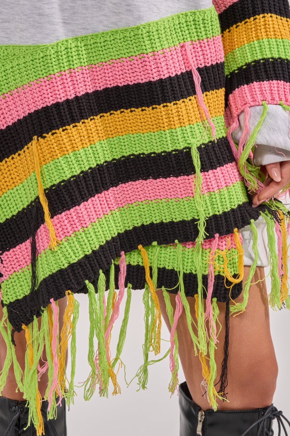 Close-up of a person wearing the MOST LOVED SWEATSHIRT MINI DRESS featuring black, green, yellow, and pink horizontal stripes with long fringe at the hem, styled boldly with shiny black boots for a standout streetwear look. 10