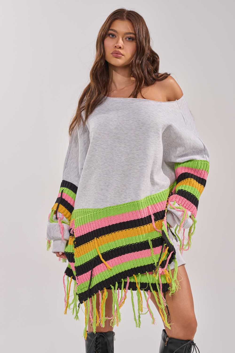 A woman stands against a plain background wearing the MOST LOVED SWEATSHIRT MINI DRESS, an oversized crochet sweater dress with neon green, pink, yellow, and black fringe on the sleeves and hem. Its loose, off-shoulder fit gives a playful streetwear vibe. 1