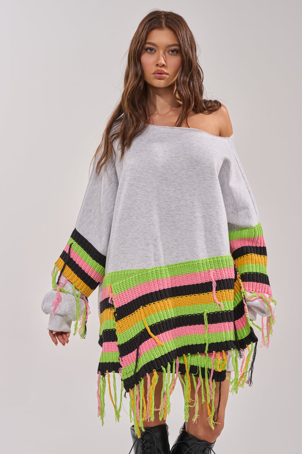 A woman stands facing forward in the MOST LOVED SWEATSHIRT MINI DRESS, an oversized crochet piece with green, pink, yellow, and black striped fringe at the hem and sleeves. She pairs it with black boots for a bold streetwear vibe. 2