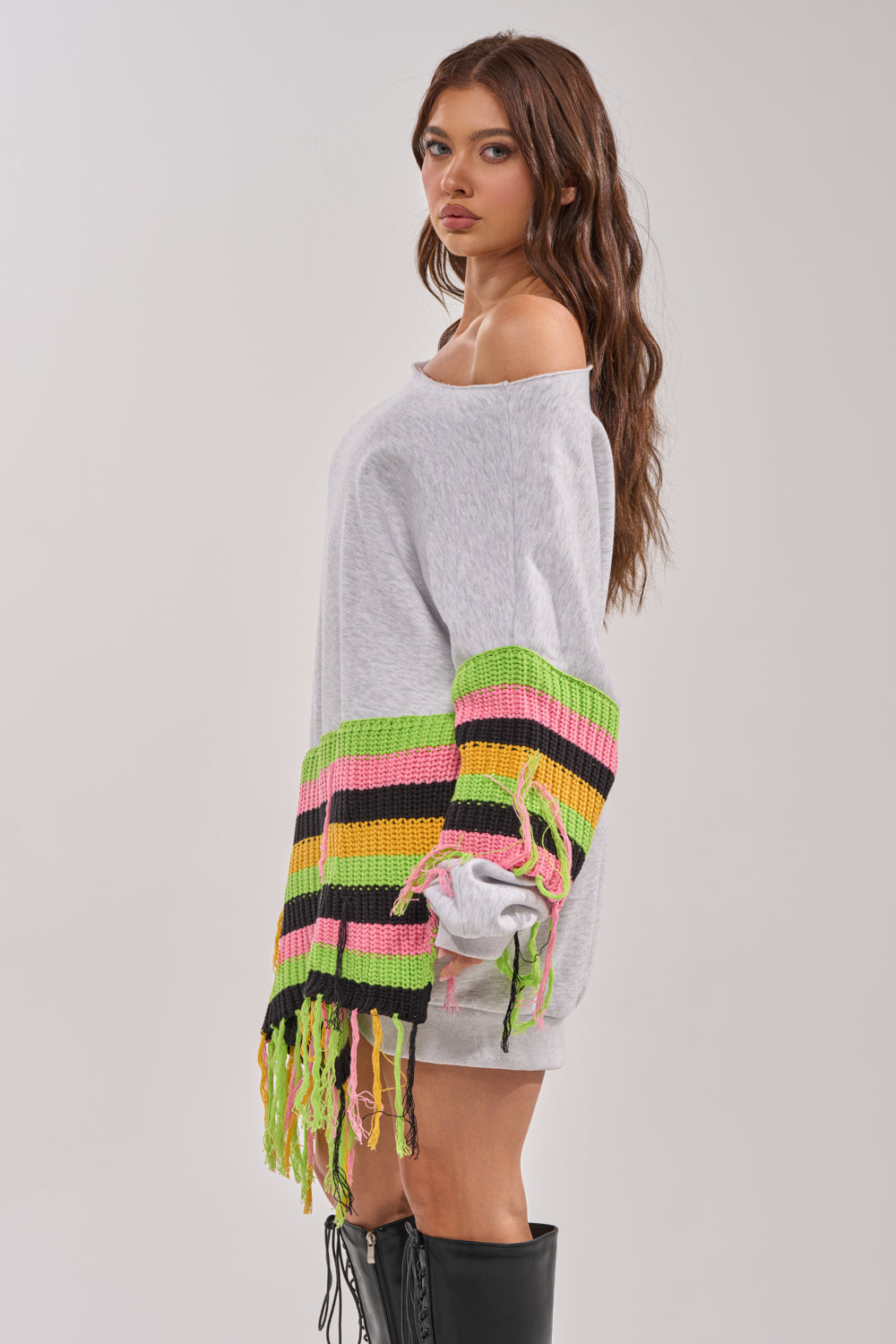 Woman in the MOST LOVED SWEATSHIRT MINI DRESS, a loose, off-shoulder mini dress with colorful crochet fringe sleeves in green, yellow, pink, and black stripes, styled sideways with knee-high black boots. 3