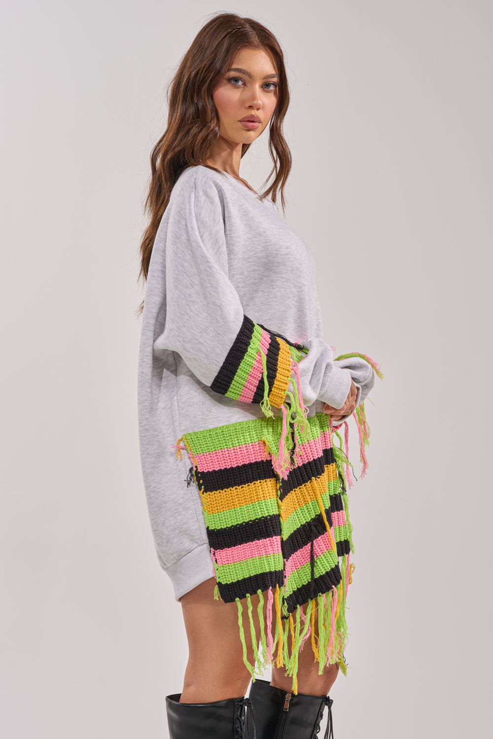 A woman with long brown hair wears the MOST LOVED SWEATSHIRT MINI DRESS—an oversized grey sweater dress with colorful knit panels and fringe—paired with black knee-high boots, standing against a plain background. 4