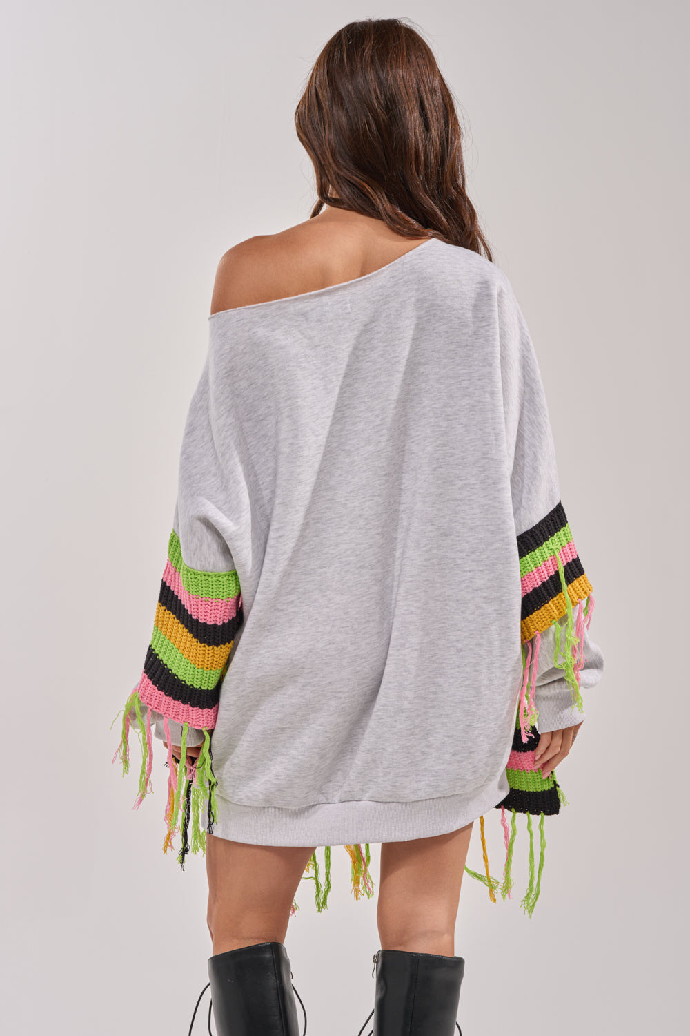 A woman stands with her back to the camera, wearing the MOST LOVED SWEATSHIRT MINI DRESS with colorful fringed sleeves. Her long brown hair flows down as she pairs the look with tall black boots for effortless streetwear style. 5