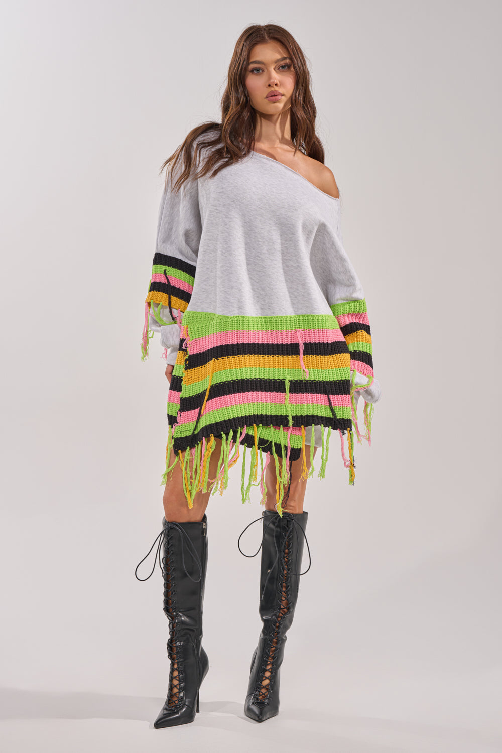 A woman wears the MOST LOVED SWEATSHIRT MINI DRESS featuring neon green, pink, and black stripes with crochet fringe, paired with knee-high black lace-up boots, standing against a plain background. 6