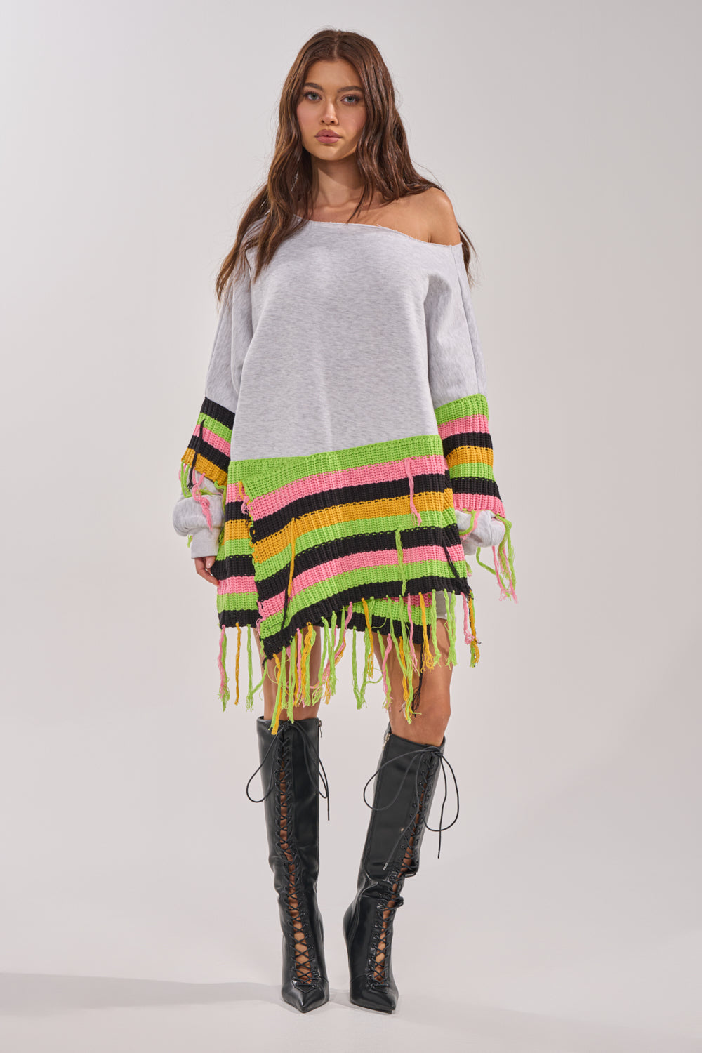 A woman wearing the MOST LOVED SWEATSHIRT MINI DRESS, featuring colorful stripes and crochet fringe, pairs it with black lace-up knee-high boots and stands against a plain light background. 7