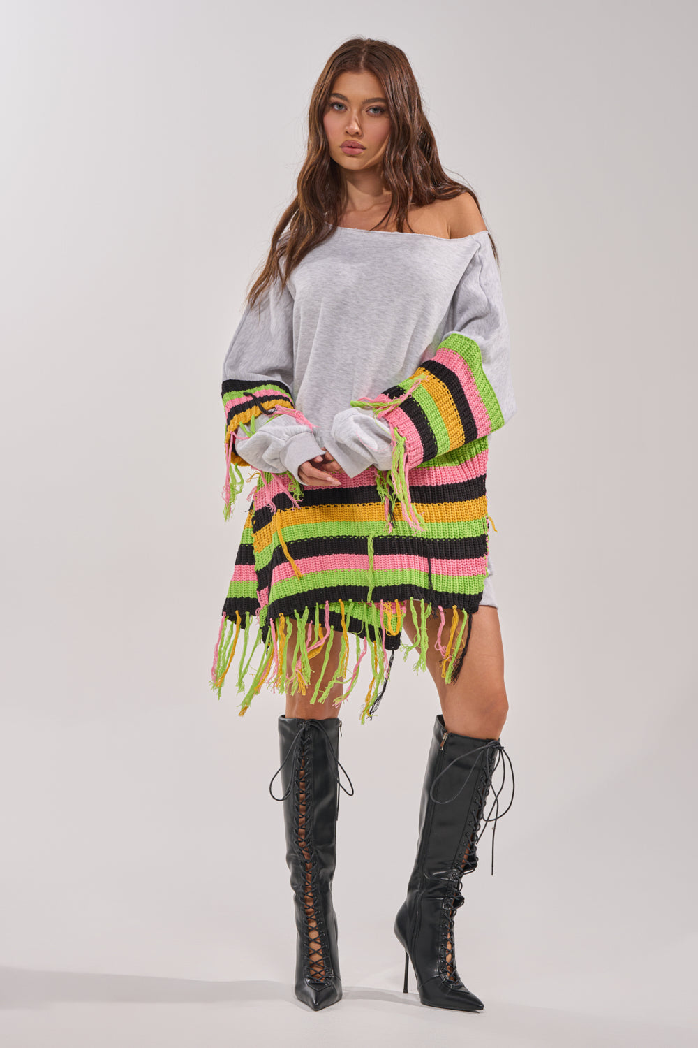A woman with long brown hair wears the MOST LOVED SWEATSHIRT MINI DRESS, an oversized gray streetwear piece featuring colorful striped crochet fringe details, paired with black lace-up knee-high boots against a plain light background. 8