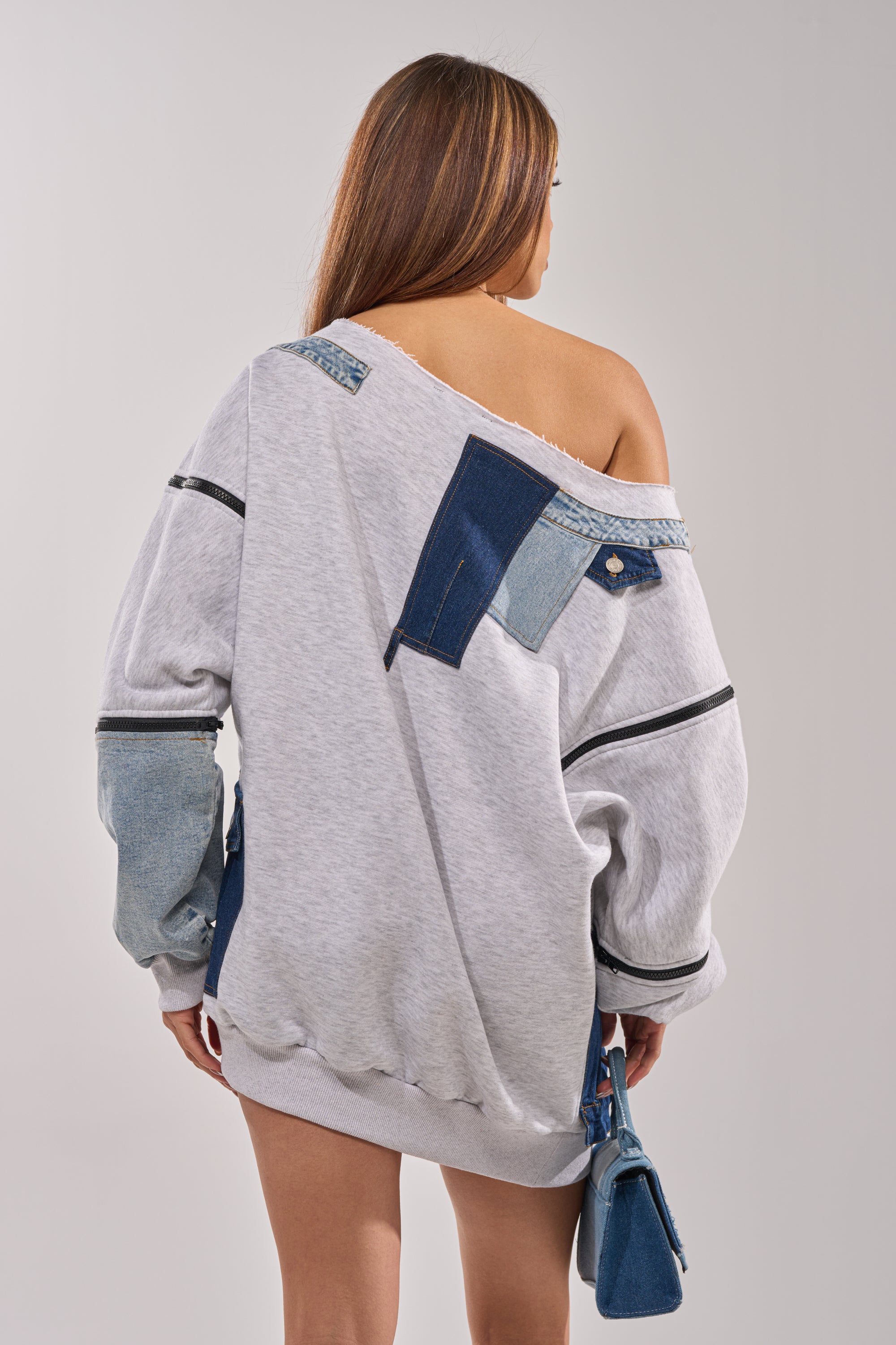 A woman with long hair, facing away, wears the ADIRA MINI SWEATER DRESS—an off-the-shoulder gray mini sweater dress with denim patches—holding a small denim bag. The relaxed fit reveals her upper back for an effortlessly chic look. 9