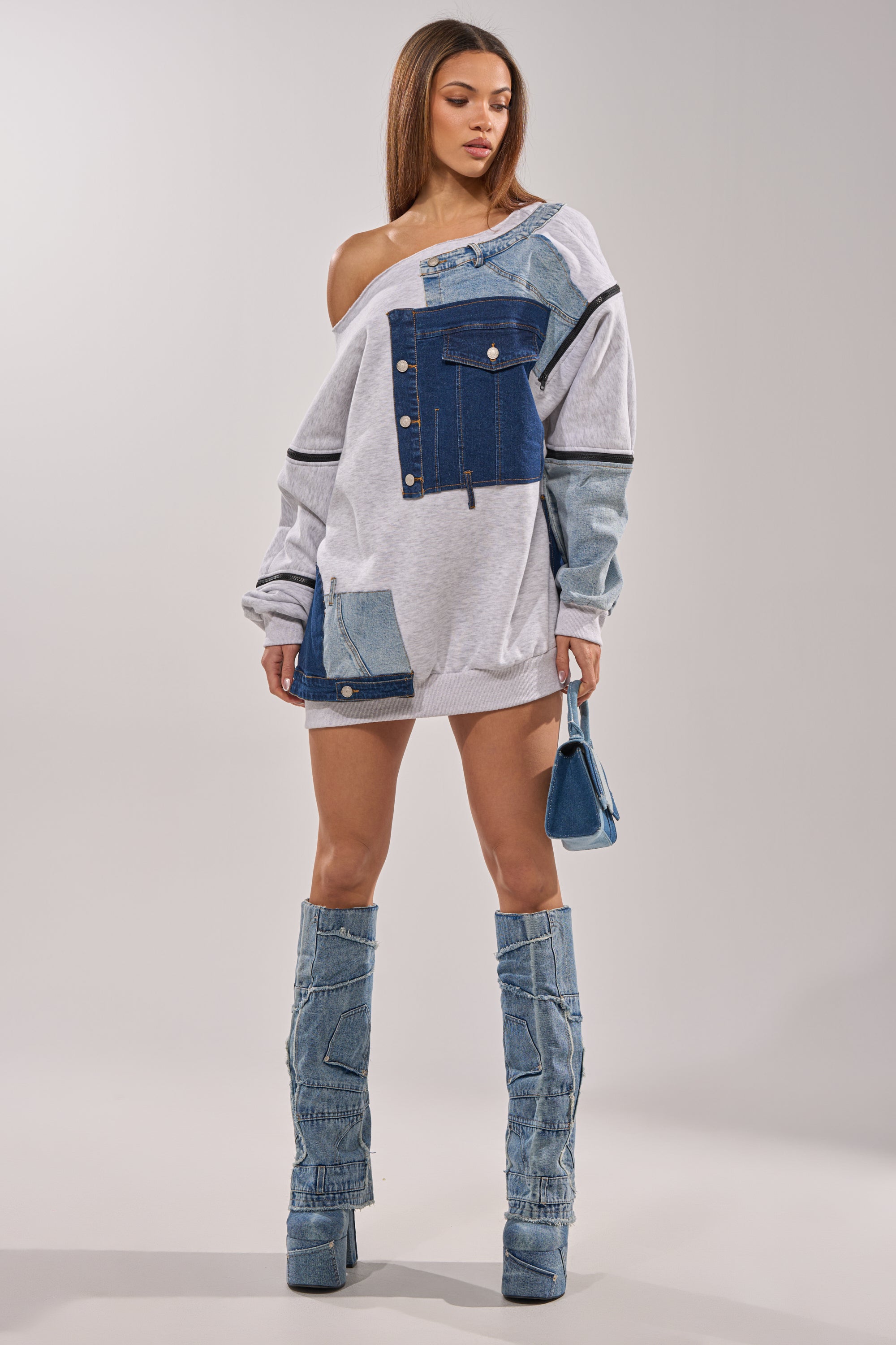 A woman wears the ADIRA MINI SWEATER DRESS, an off-the-shoulder style blending light gray fabric with denim panels and metallic buttons. She pairs it with tall denim boots and carries a small blue purse for a chic look. 11