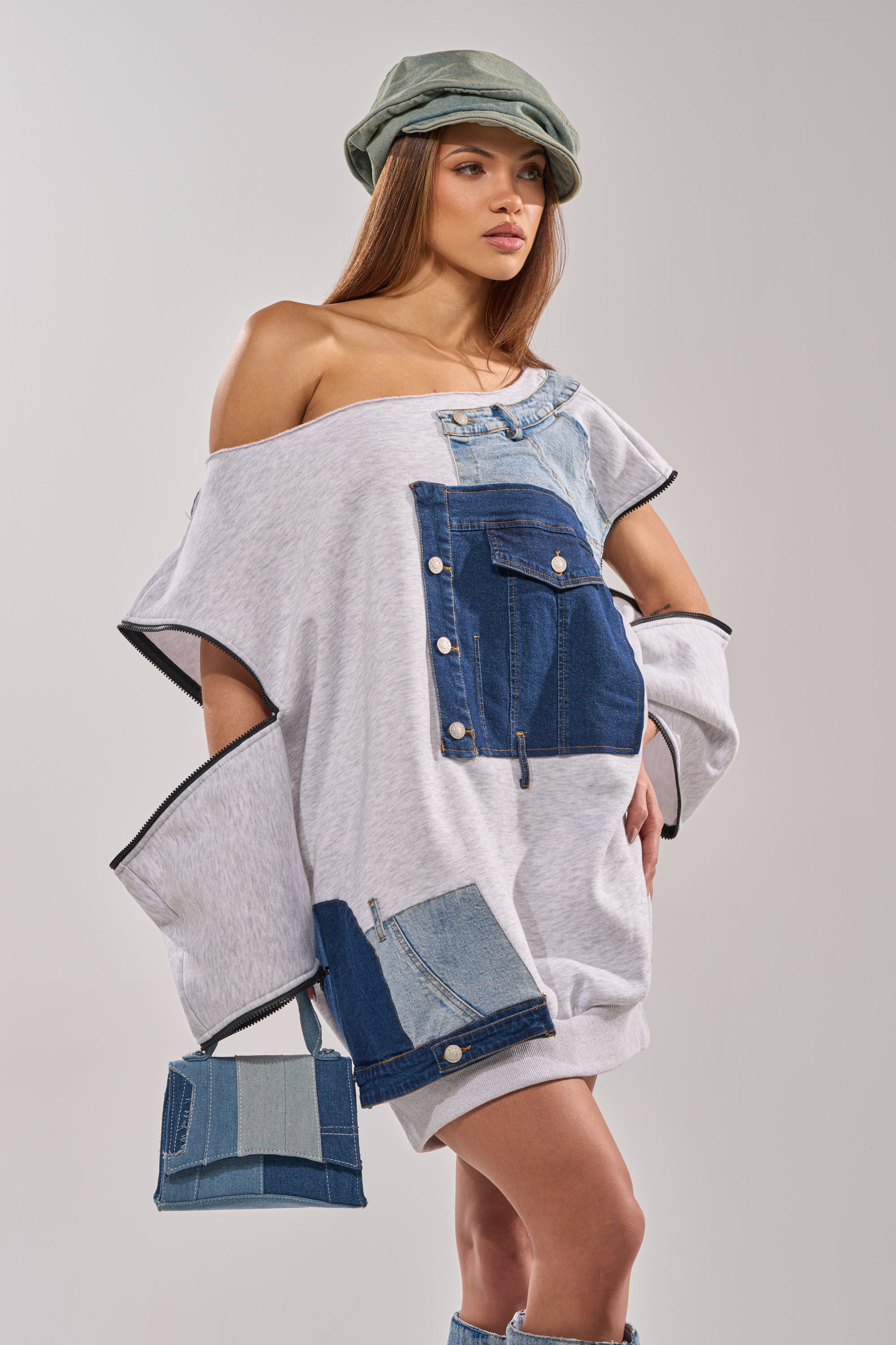 A woman poses confidently in the ADIRA MINI SWEATER DRESS with a green cap, holding a small patchwork denim bag against a plain background. 0