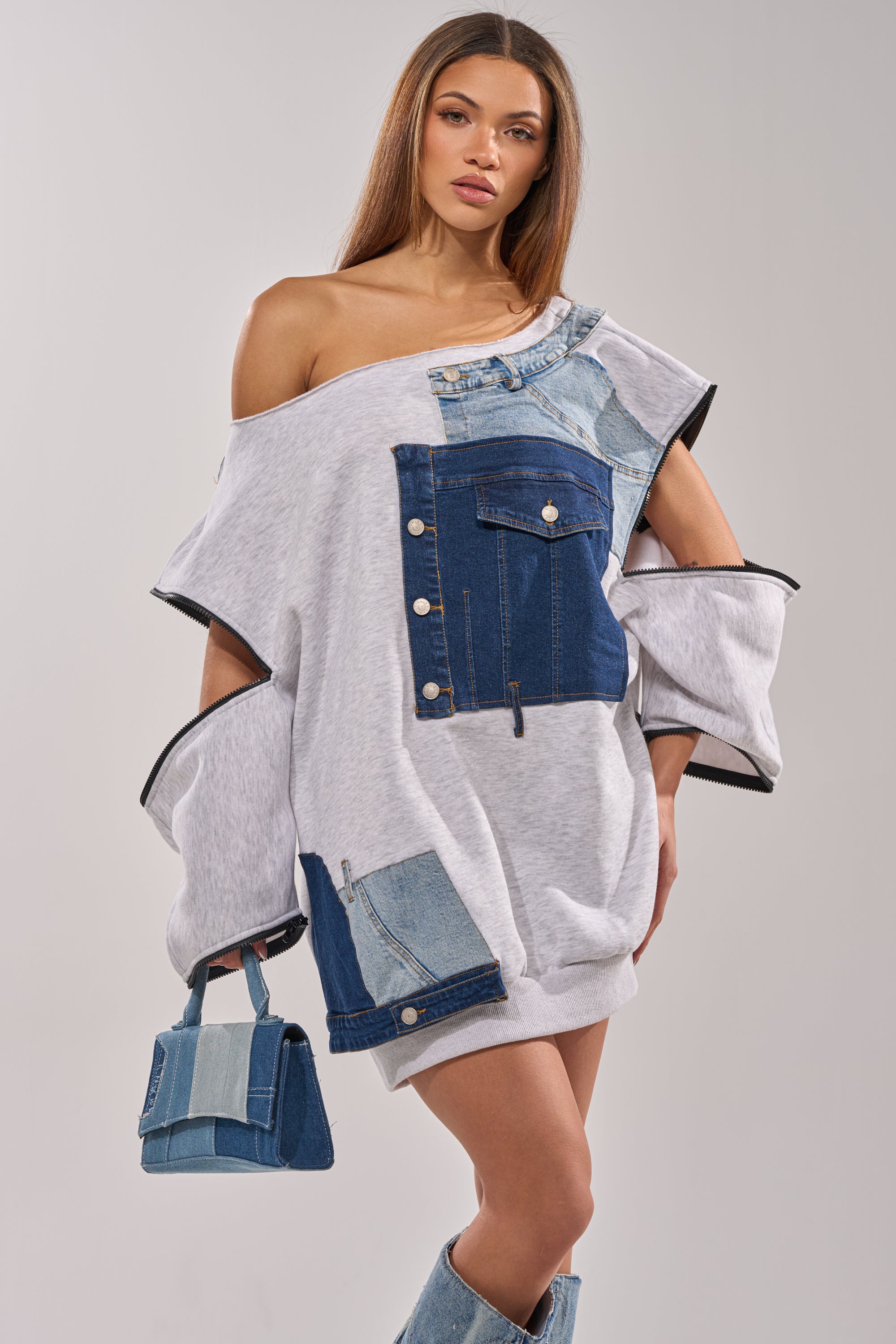A woman poses in the oversized gray ADIRA MINI SWEATER DRESS with denim patchwork, styled with matching denim boots and a small denim handbag. Her straight hair and the neutral background complete the chic look. 1