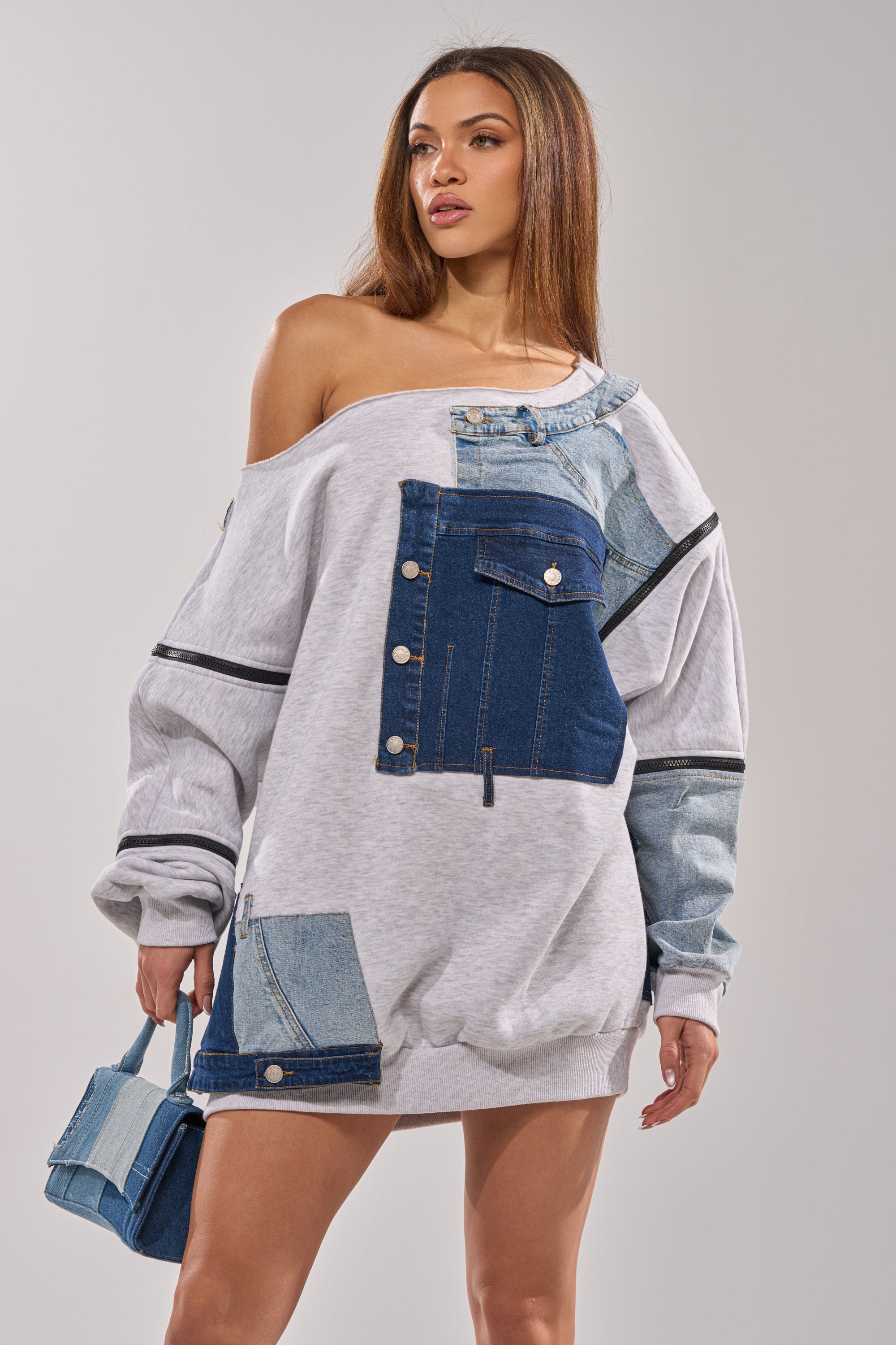 A woman models the ADIRA MINI SWEATER DRESS, an oversized off-the-shoulder mini with denim patchwork details, paired with a matching mini handbag against a plain light background. 2