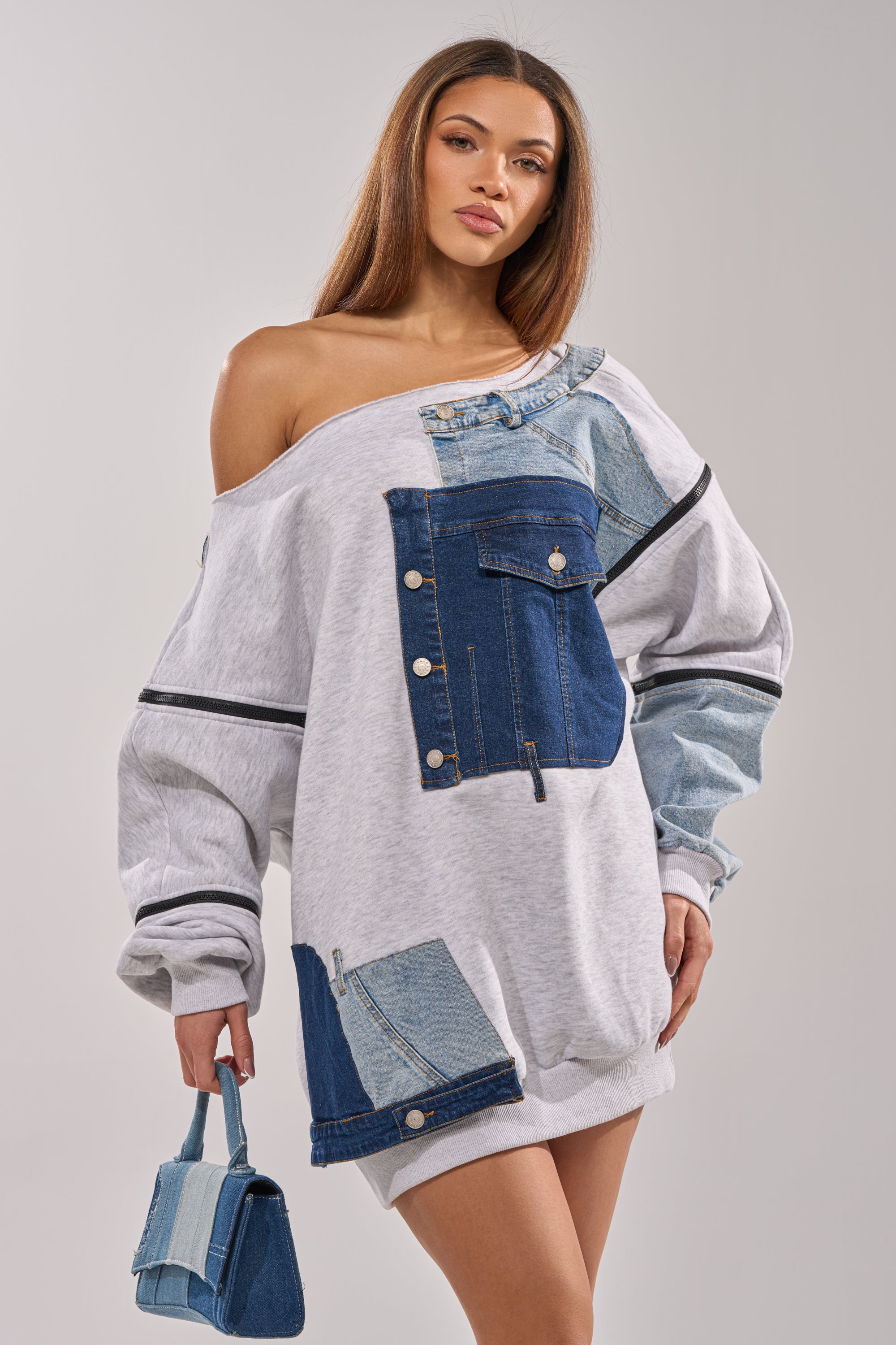 A woman poses against a plain background wearing the ADIRA MINI SWEATER DRESS—an oversized, off-the-shoulder mini dress with denim patchwork details—and holds a matching small denim patch handbag. 3