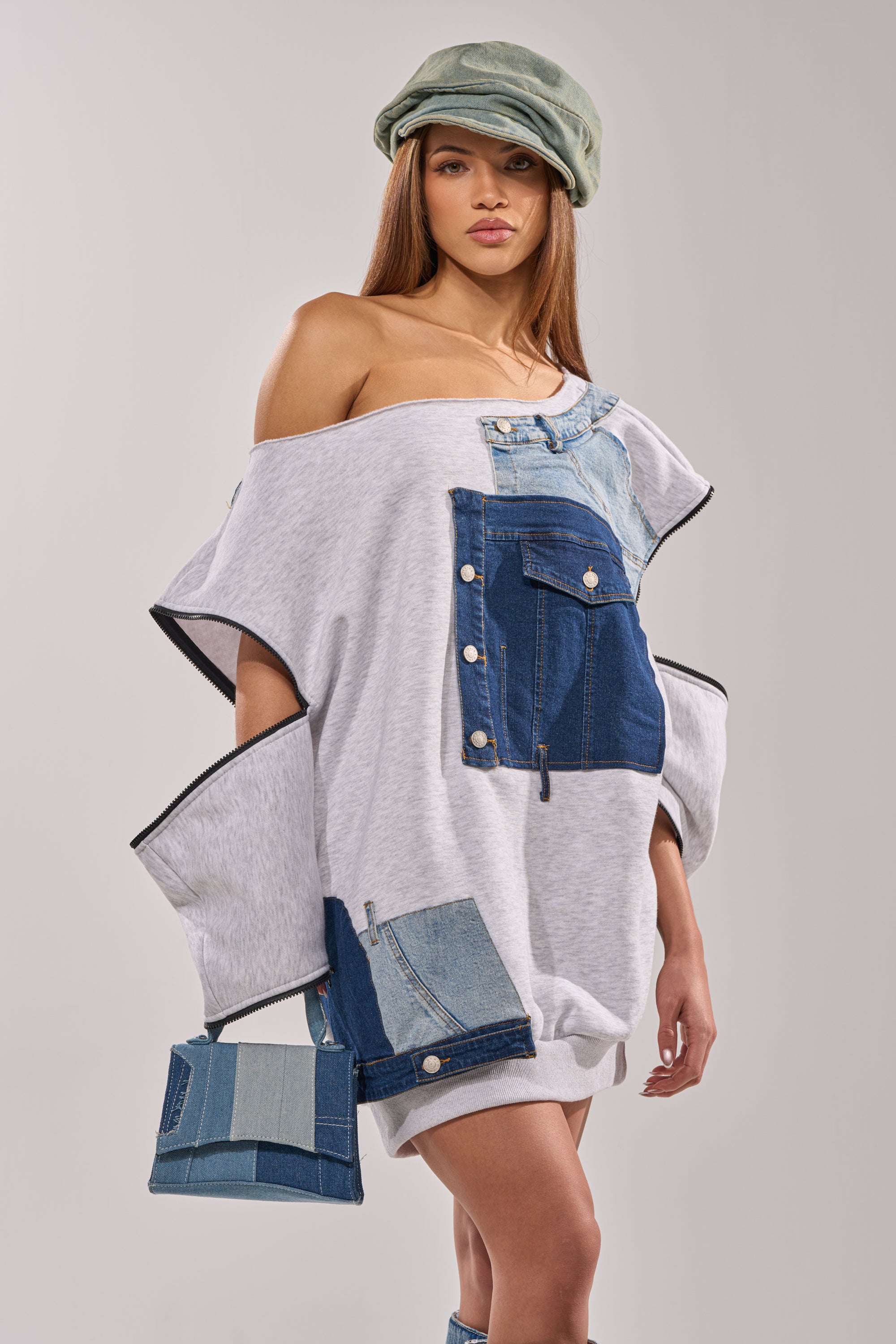 A woman wears the oversized ADIRA MINI SWEATER DRESS with an off-the-shoulder gray denim patchwork design, holding a matching denim handbag. She has long hair and a light green cap against a plain, light background. 6