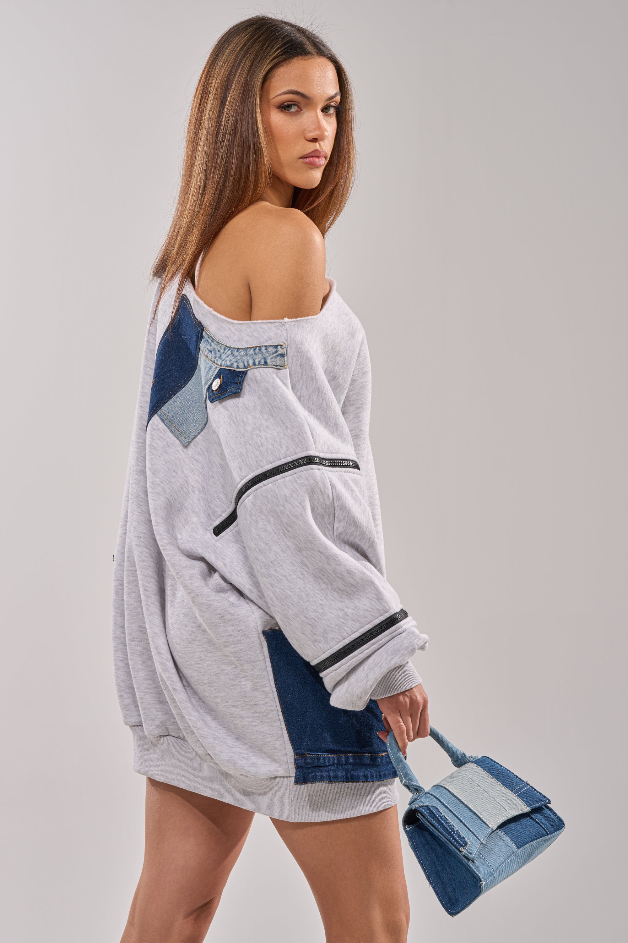 Wearing the ADIRA MINI SWEATER DRESS with blue denim patches, a woman with straight brown hair holds a small blue denim handbag and looks over her shoulder against a plain background. 8