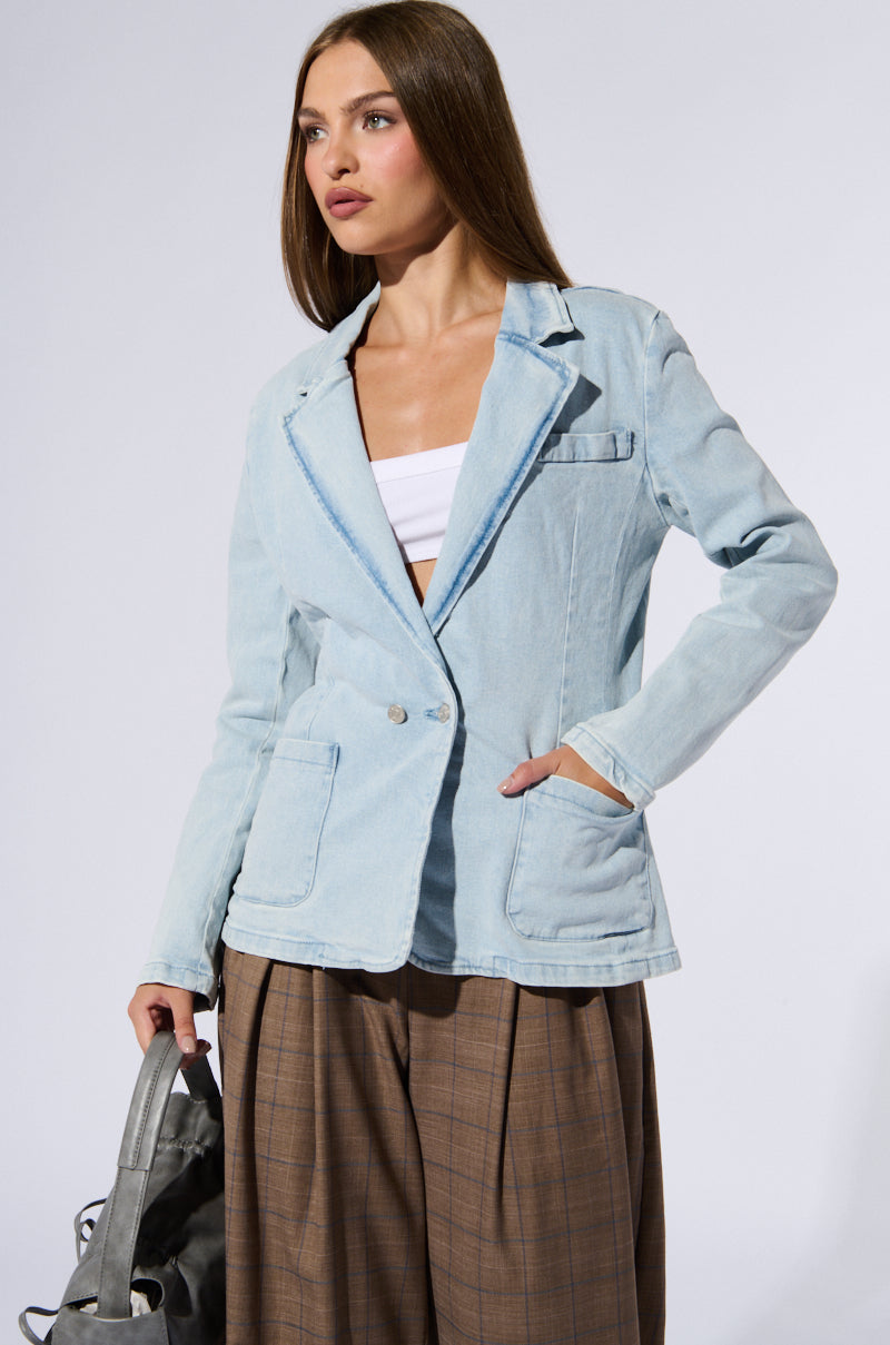 The woman exudes business chic in her light blue ROBYN DENIM BLAZER over a white top and brown plaid wide-leg pants. Holding a gray bag in her left hand and with her right hand in her blazer pocket, she gazes off to the side against a plain background.
