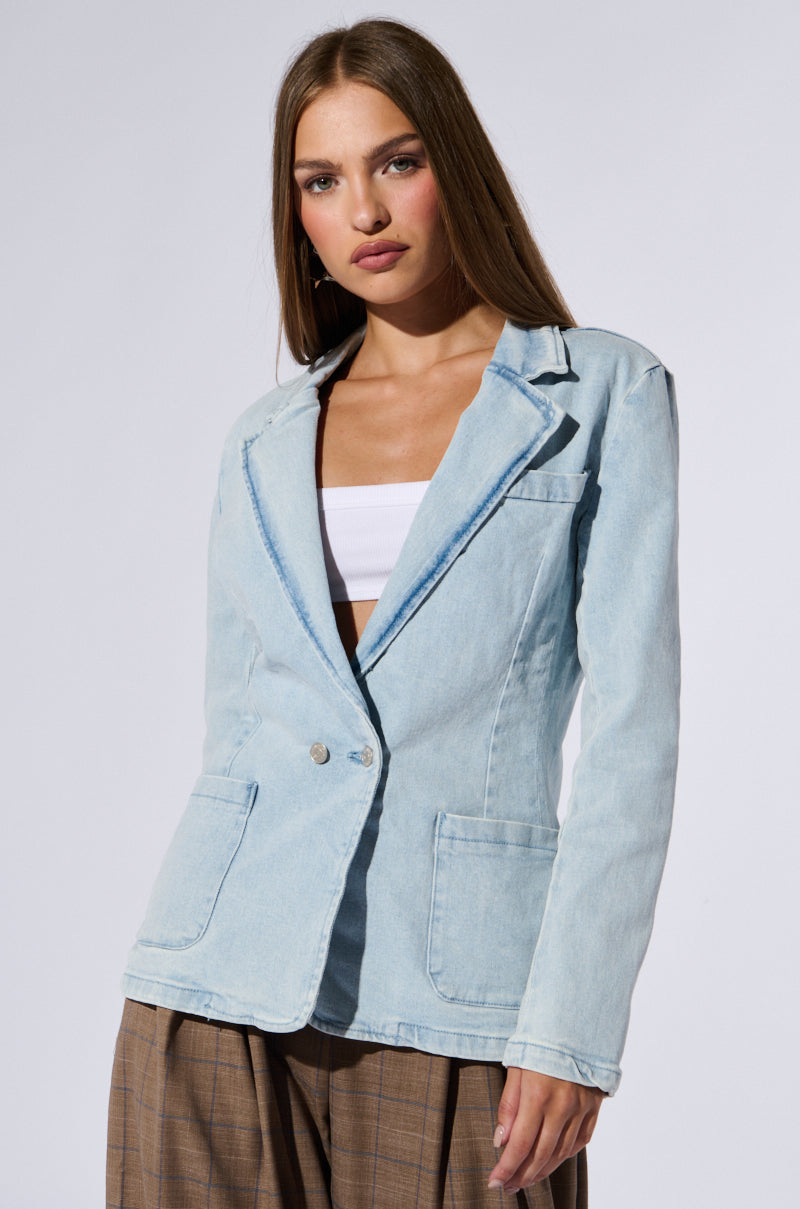 A woman with long brown hair stands against a plain background, exuding business chic. She is wearing a light blue ROBYN DENIM BLAZER over a white top and brown plaid pants. With its relaxed fit, she looks directly at the camera with a neutral expression.