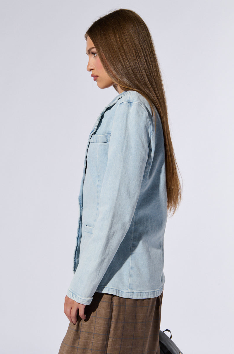 A woman with long, straight brown hair is standing in profile against a plain background. She is wearing a ROBYN DENIM BLAZER in light blue over a brown plaid skirt, exuding an understated business chic vibe. The image focuses on her left side, highlighting the relaxed fit of her outfit.