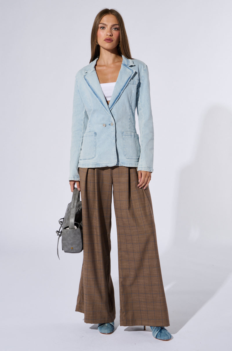 A woman is standing against a plain background. She is wearing a ROBYN DENIM BLAZER over a white top, paired with brown wide-leg plaid pants. She holds a gray handbag in one hand and wears blue shoes. Her long hair is parted in the middle as she gazes forward, exuding business chic style.