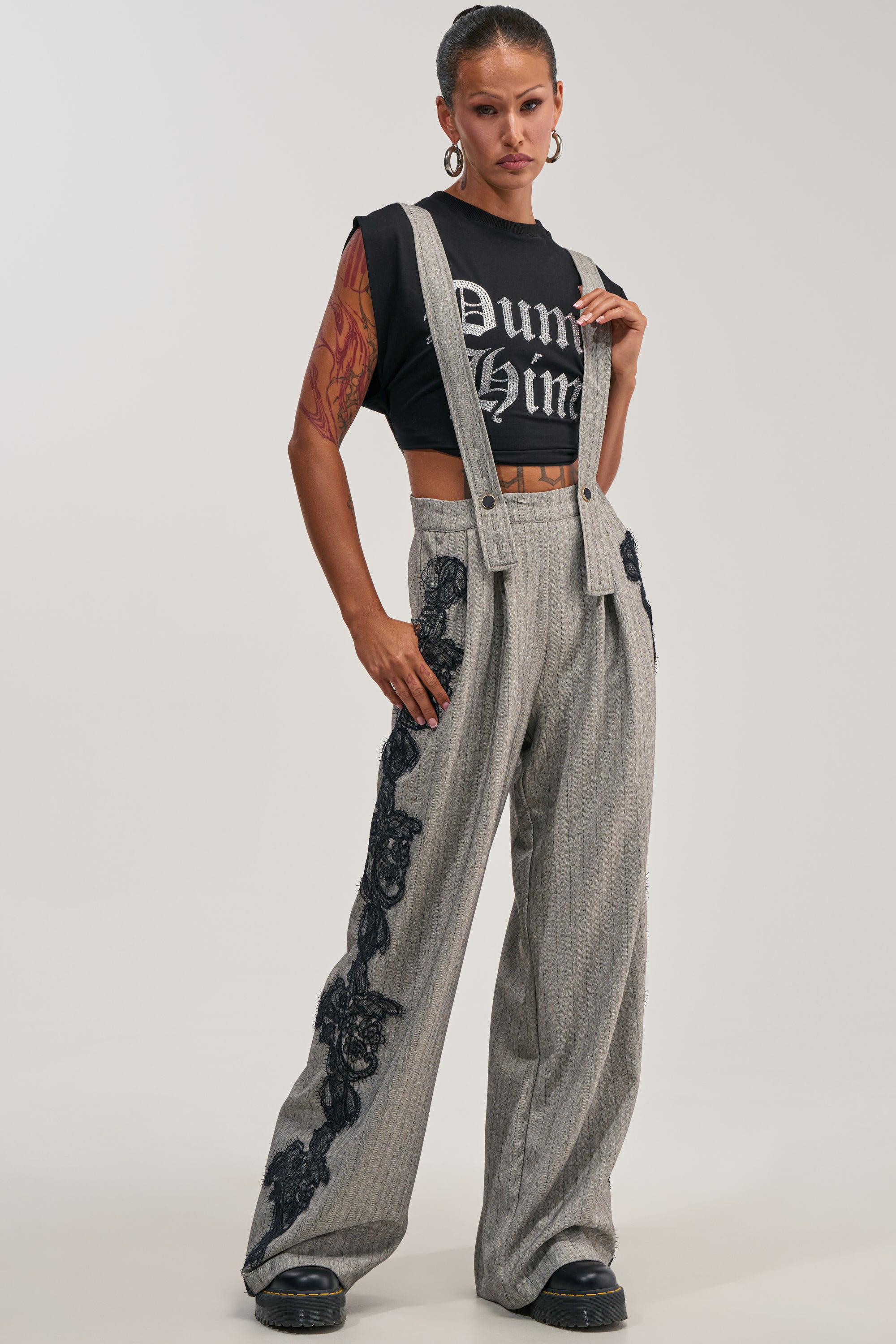 Confidently standing against a plain white background, she showcases contemporary styling and statement workwear in the FOREVER FLAWLESS PINSTRIPE OVERALLS—grey, wide-leg suspenders with black lace side detail—paired with a black graphic crop top.
