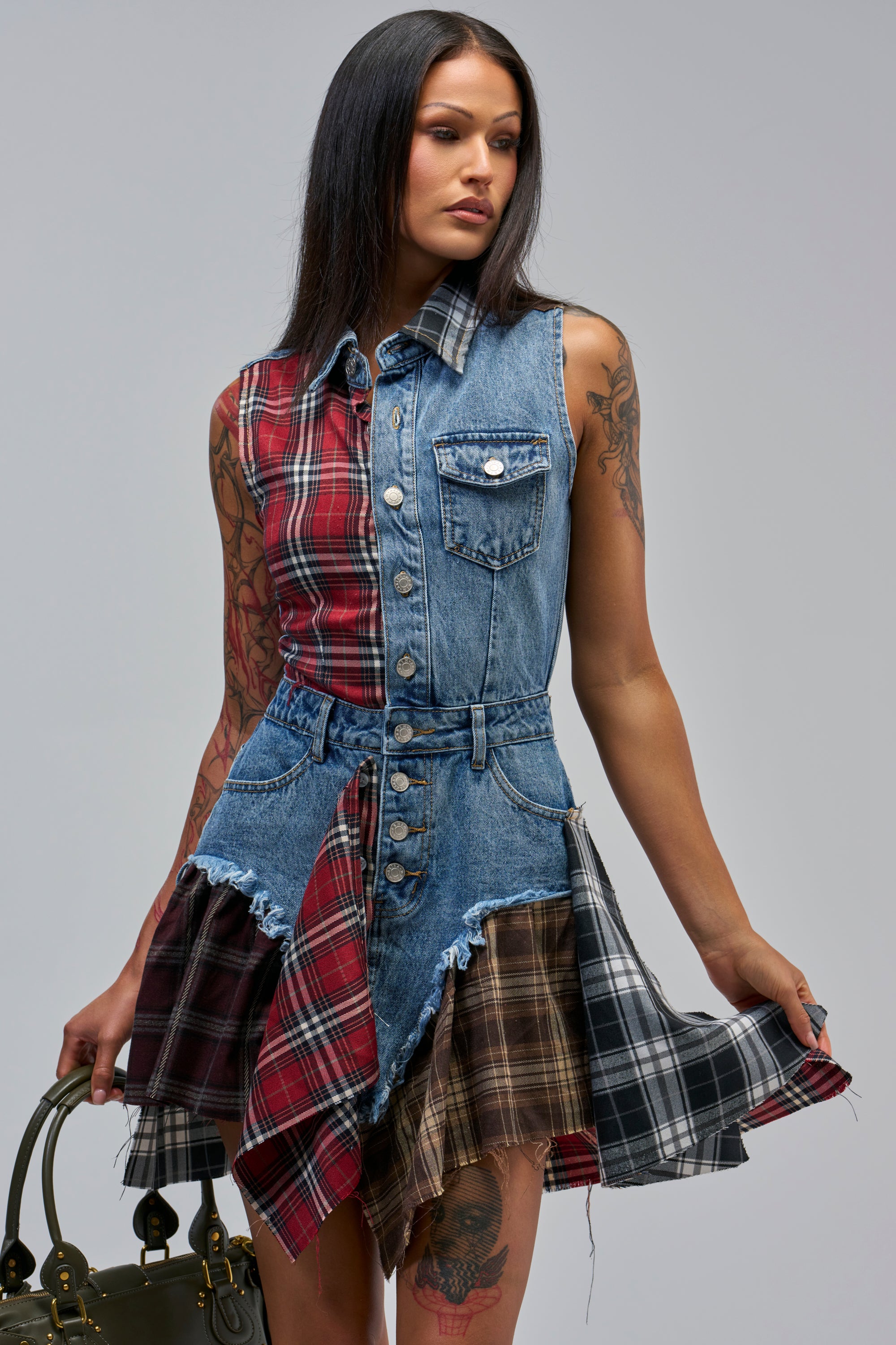 A woman with long dark hair wears the READY TO RIDE PATCHWORK DENIM MINI DRESS, featuring plaid panels. She holds her skirt, carries a handbag, and shows tattoos on her arms and legs.