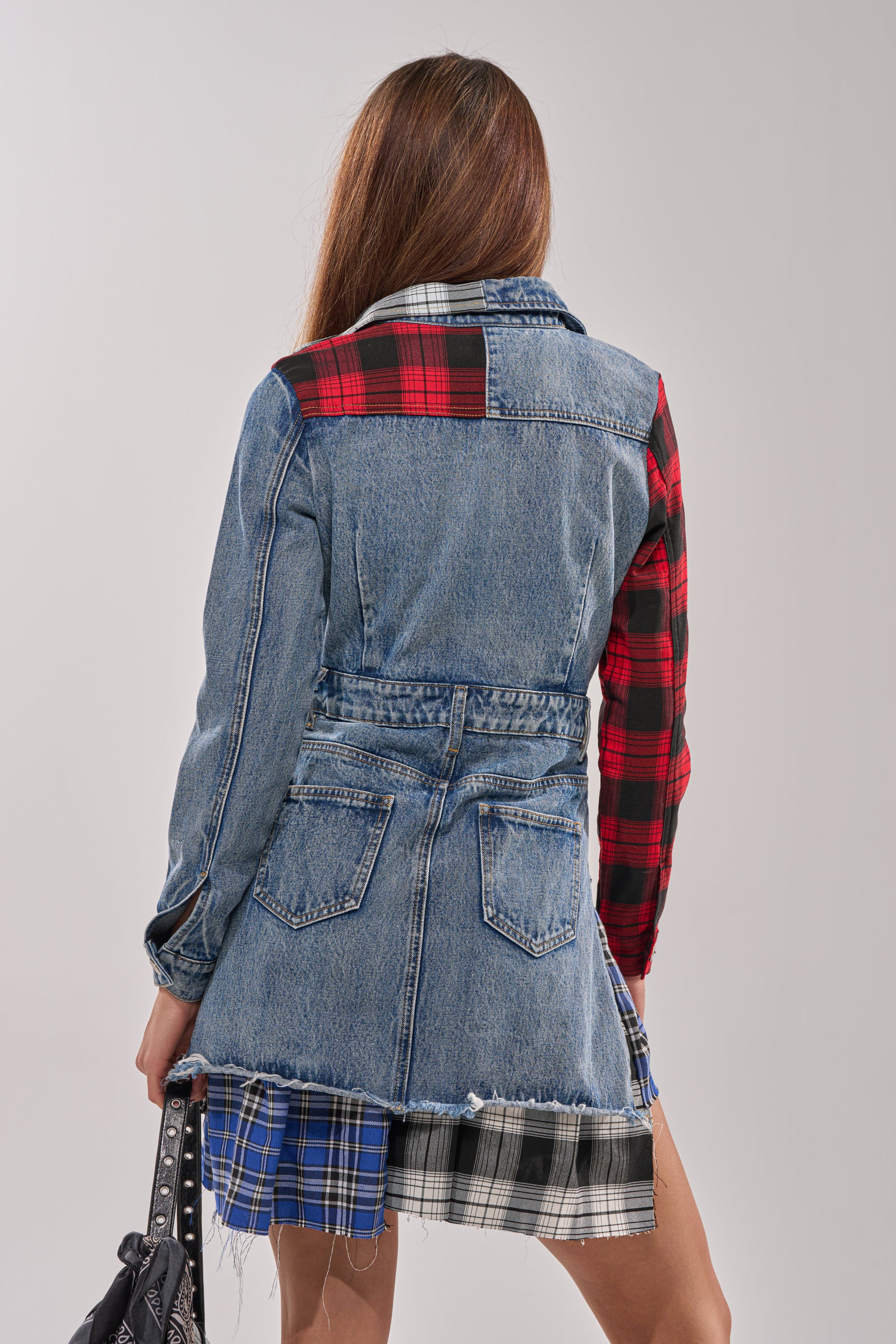 A woman with long brown hair is seen from behind wearing the PATCHWORK PLAID DENIM MINI DRESS—a punk-inspired mini dress with plaid sleeves, patchwork details, an asymmetrical hem, and a denim jean belt. She holds a studded black bag.