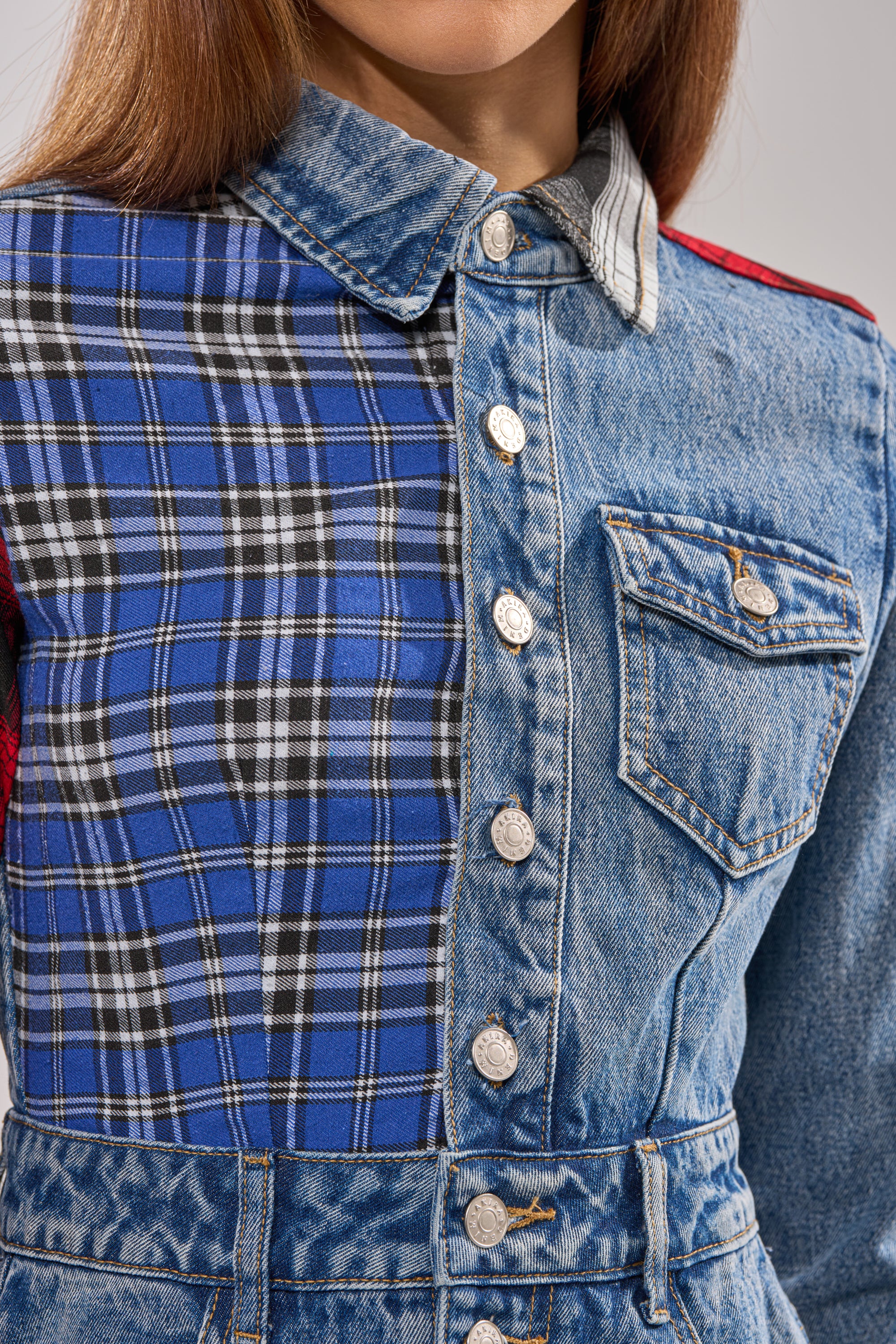 Close-up of a person wearing the PATCHWORK PLAID DENIM MINI DRESS, featuring punk-inspired silver buttons, patchwork plaid on one side, and solid blue denim with a chest pocket on the other. The face is not visible.