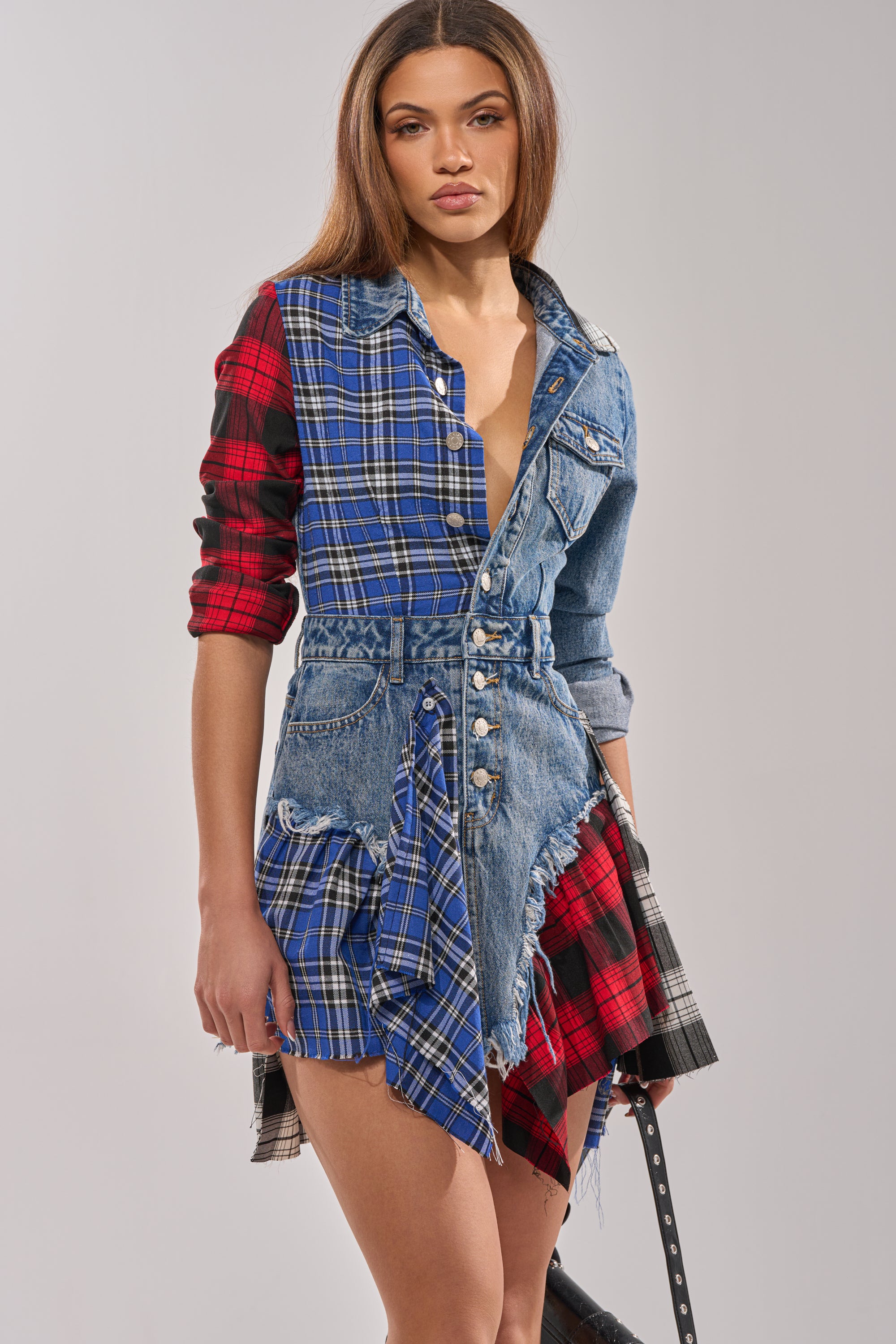 Wearing the PATCHWORK PLAID DENIM MINI DRESS with a button-down front, frayed edges, and asymmetrical hem, a woman with long straight hair poses with a neutral expression.