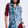 A person wearing an edgy ensemble stands confidently in the PATCHWORK PLAID DENIM MINI DRESS, featuring red and blue elements, frayed edges, and button details. They have shoulder-length black hair and a neutral expression against a plain background.