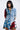 A woman models the PATCHWORK PLAID DENIM MINI DRESS, blending denim with red, blue, and black plaid patterns. The dress features a jean jacket-style top with buttons and frayed edges. Her shoulder-length black hair stands out against the white background.