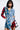 A woman stands confidently in a PATCHWORK PLAID DENIM MINI DRESS. The punk-inspired dress features multicolored plaid sections, a fringed hem, long sleeves, and button details. Her shoulder-length black hair frames her face as she casually keeps her hands in her pockets.