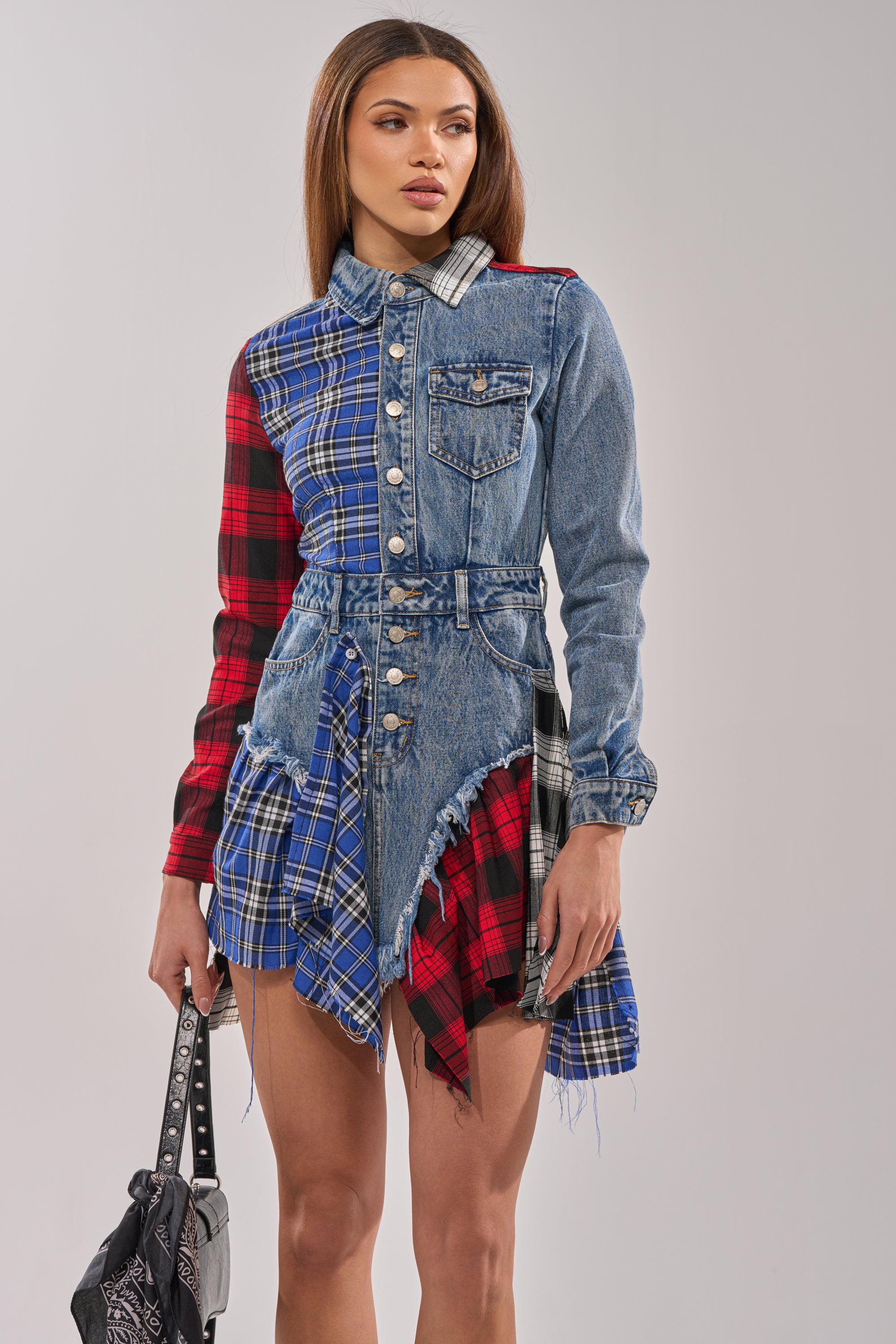 A woman models the PATCHWORK PLAID DENIM MINI DRESS—featuring denim, blue and red plaid panels with frayed edges and an asymmetrical hem—while holding a black handbag against a plain background.