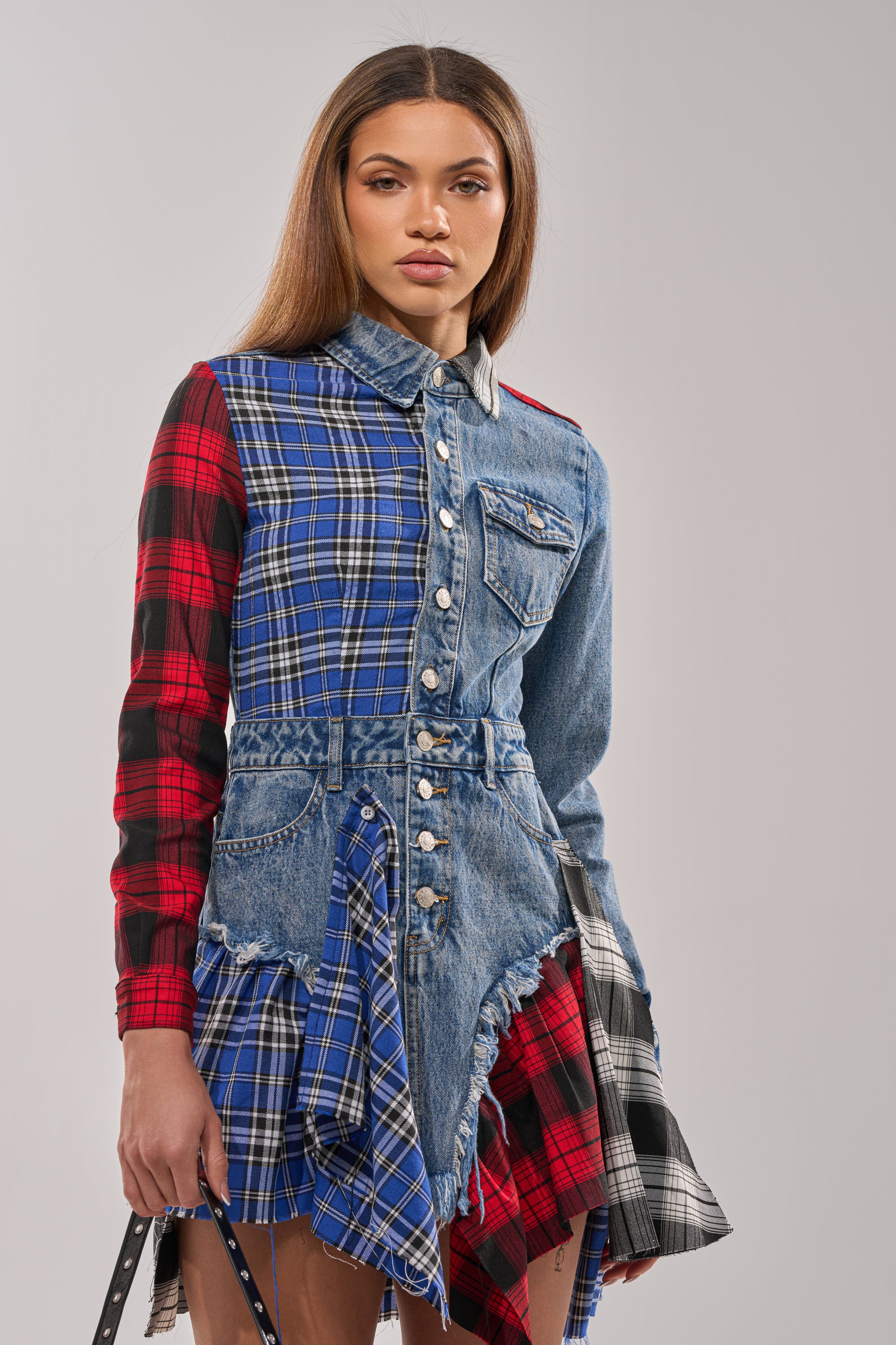 A woman stands against a plain light background wearing the PATCHWORK PLAID DENIM MINI DRESS, featuring frayed edges, a button-up front, and an asymmetrical hem for a unique punk-inspired look.