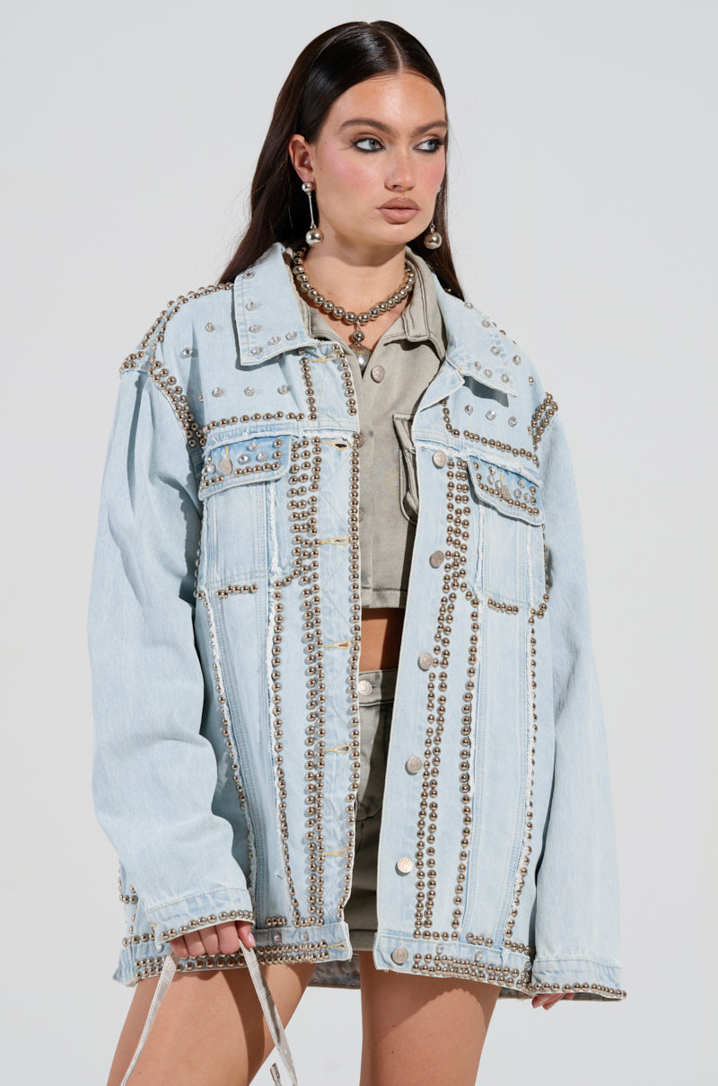 A woman with long dark hair stands in the eye-catching GAME TIME EMBELLISHED LONGLINE DENIM JACKET adorned with metal studs. She pairs it perfectly with matching studded shorts and a beige crop top, complementing her look with hoop earrings, a chain necklace, and holding a white bag in her left hand.