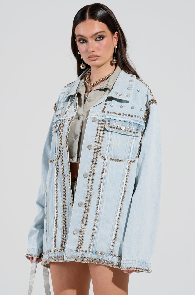 A person wearing the GAME TIME EMBELLISHED LONGLINE DENIM JACKET, adorned with silver studs, is paired with a light gray shirt and large hoop earrings, standing against a plain background. Their dark hair falls straight past their shoulders.