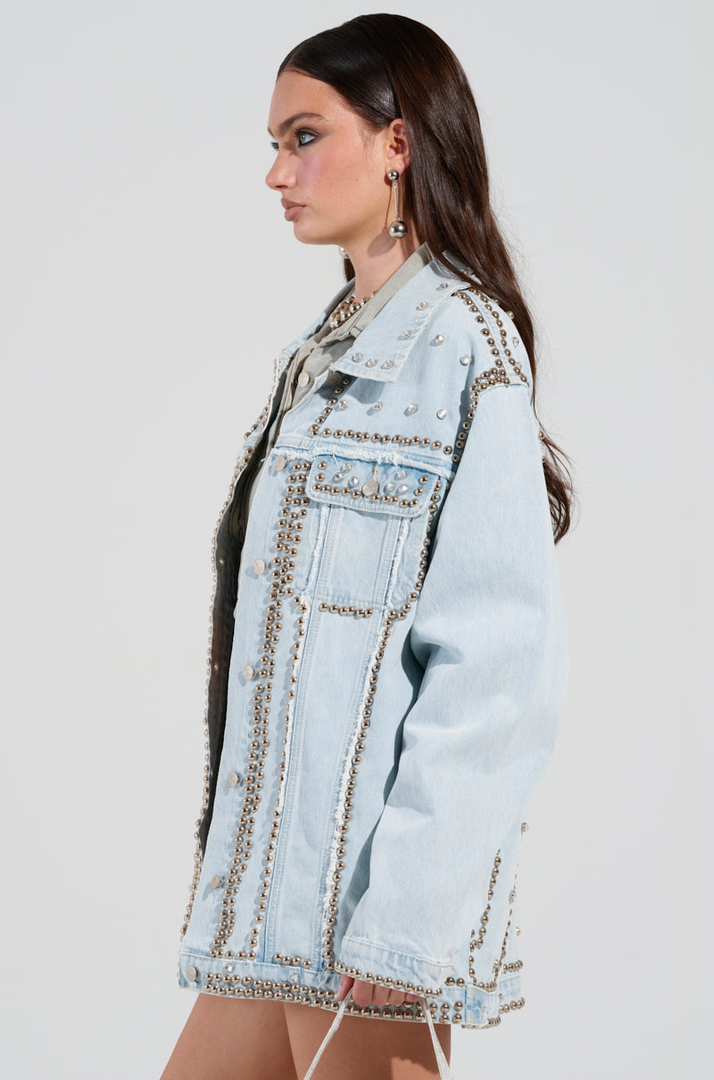 A person with long, dark hair is standing in profile against a plain background, wearing the GAME TIME EMBELLISHED LONGLINE DENIM JACKET. This oversized light blue jacket is adorned with silver studs.