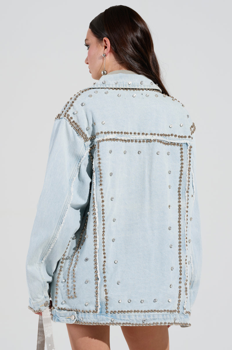 A person with long dark hair is wearing the GAME TIME EMBELLISHED LONGLINE DENIM JACKET, featuring a light blue hue adorned with silver studs. They are turned away from the camera, highlighting the decorative back of the jacket against a plain background.