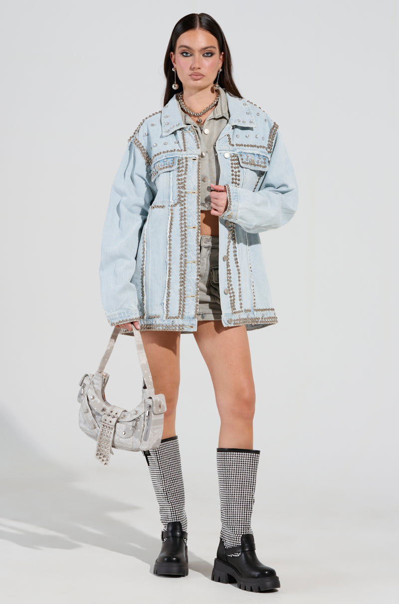 A woman poses elegantly in a chic ensemble, featuring the GAME TIME EMBELLISHED LONGLINE DENIM JACKET adorned with studs, a gray outfit beneath it, thigh-high rhinestone boots, and a coordinating studded handbag. Her hair flows long and straight.