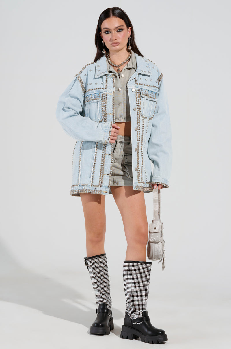 A person stands confidently in a light blue GAME TIME EMBELLISHED LONGLINE DENIM JACKET over a gray outfit. They hold a white bag and wear knee-high socks with black chunky boots. Their hair is long and dark, beautifully adorned with elegant jewelry.