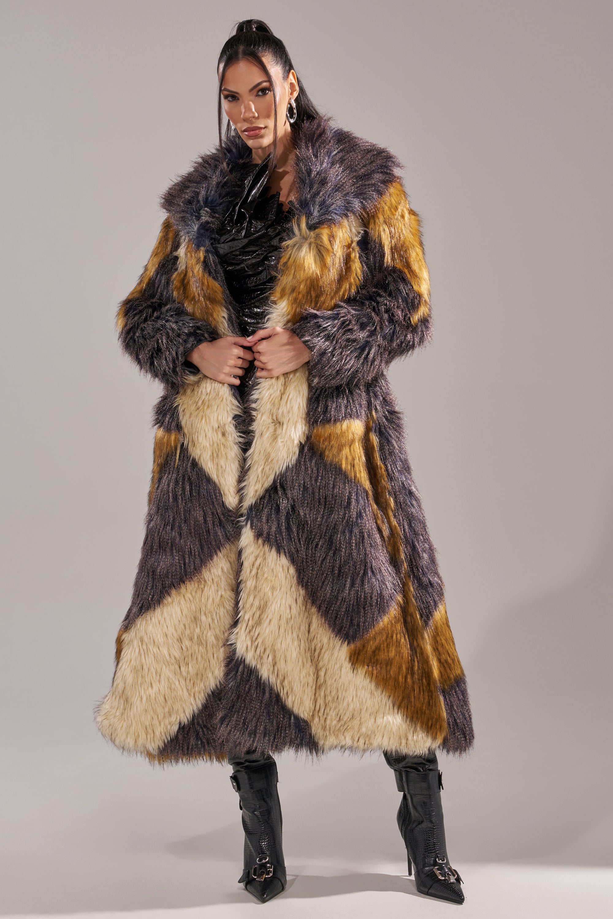 A woman stands confidently in the NOTRE DAME FAUX FUR COAT, featuring a long patchwork design in yellow, brown, and cream over a black outfit with heeled boots against a plain light gray background.