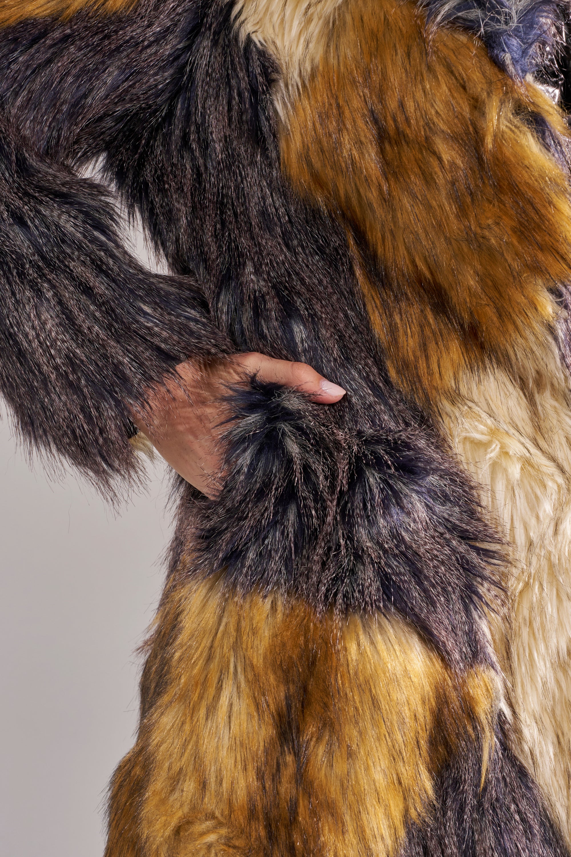 A close-up shows a hand in the pocket of the NOTRE DAME FAUX FUR COAT, featuring a multicolored patchwork design in brown, black, and cream. Its soft plush texture and luxe street style make it a standout piece.