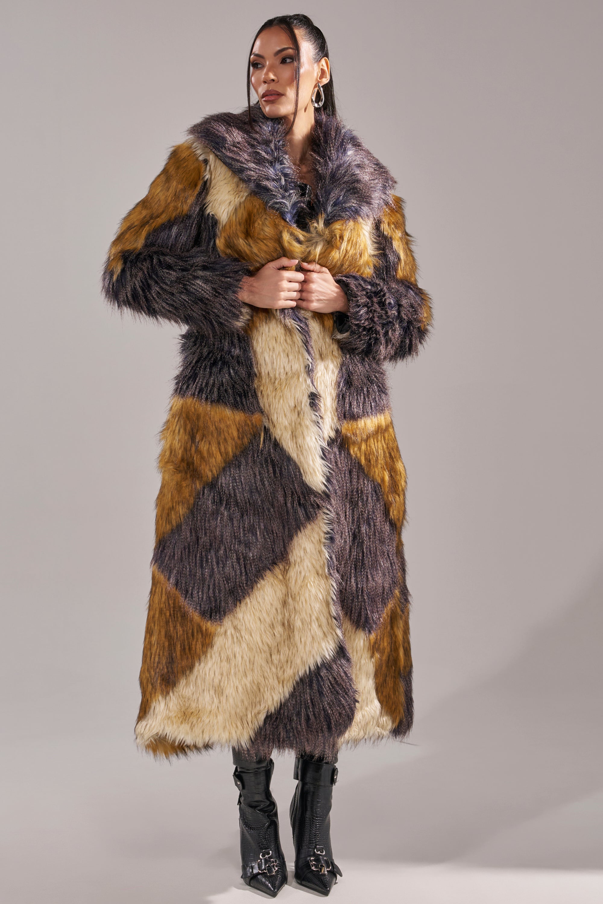 A woman poses confidently in a studio, wearing the NOTRE DAME FAUX FUR COAT featuring a patchwork design in brown, beige, and dark purple. Paired with black boots against a neutral backdrop, she captures the essence of luxury street style.