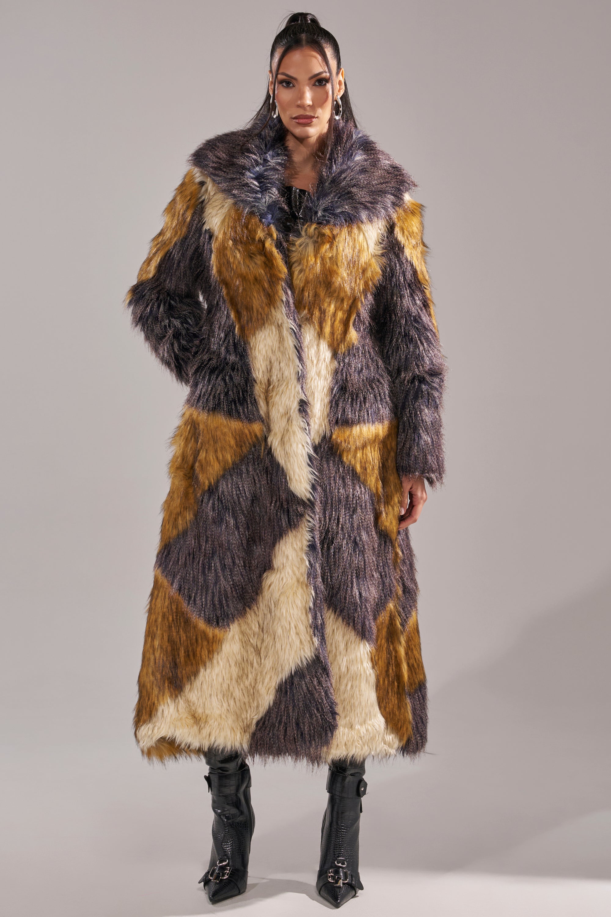 A person models the NOTRE DAME FAUX FUR COAT, a long patchwork design in brown, cream, and gray tones, styled with black heeled boots for a luxe street style look against a neutral backdrop.